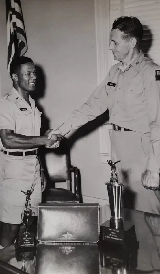 Black-and-white photo: Johnson in uniform shaking hands with an older officer; on the table, two trophies for the 3-meter springboard diving event at Fort Totten