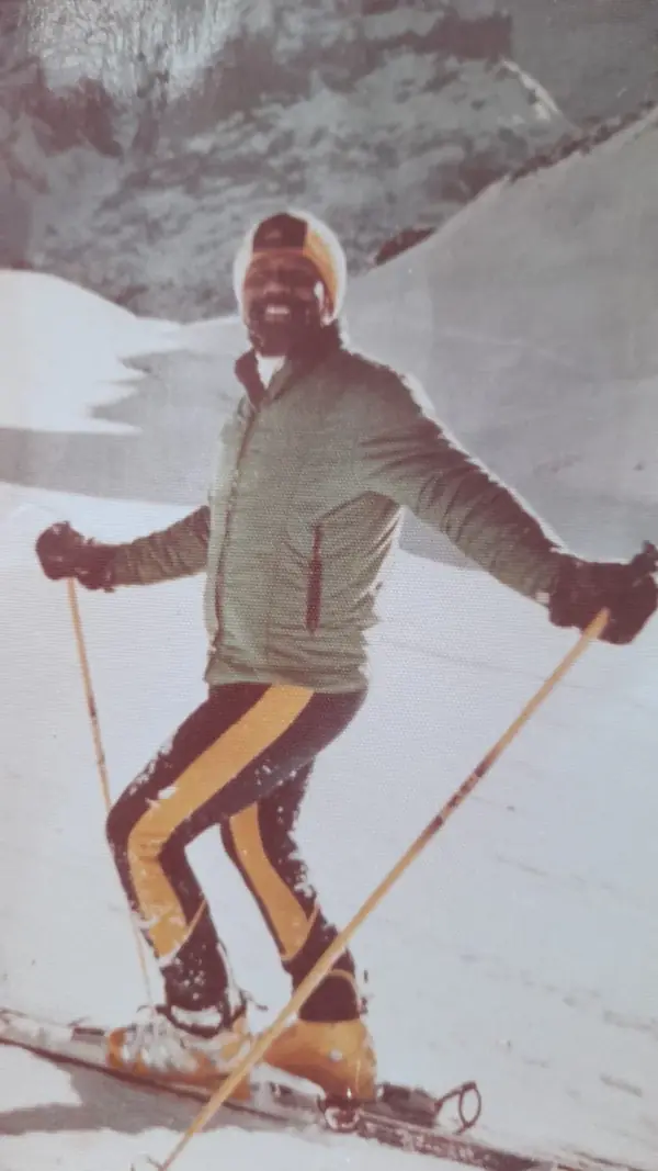 Color photo from the 1970s: Johnson laughing on skis, in a green jacket and striped ski pants with a wool cap
