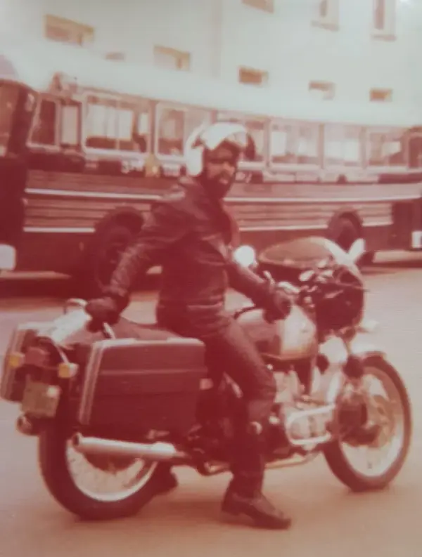 Color photo from the 1970s: Johnson with a beard, leather jacket, helmet and sunglasses on a touring motorcycle with luggage cases, a city bus in the background