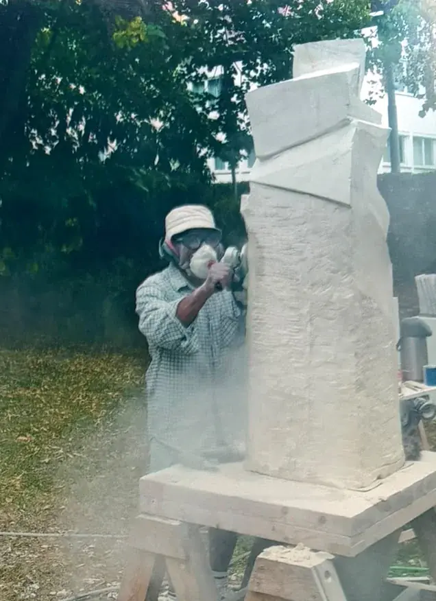 Johnson in a hat, goggles, dust mask and ear protection grinding a tall pale stone stele
