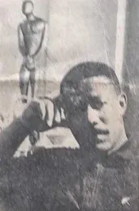 Newspaper photo from 1968: Ronald E. Johnson, his hand on his forehead, in front of a standing wooden figure